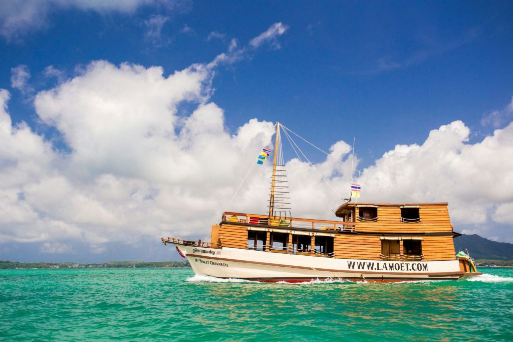 "WWW.LAMOET.COM" 웹사이트가 있는 목조 투어 보트는 부분적으로 흐린 푸른 하늘 아래 맑은 청록색 물 위에서 항해합니다.