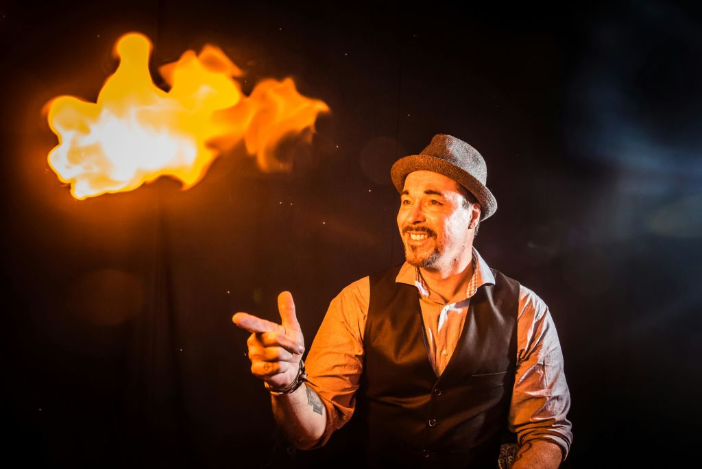 조끼와 모자를 쓴 남성이 조명이 어두운 환경에서 불꽃을 향해 손짓하며 미소를 짓고 있습니다.