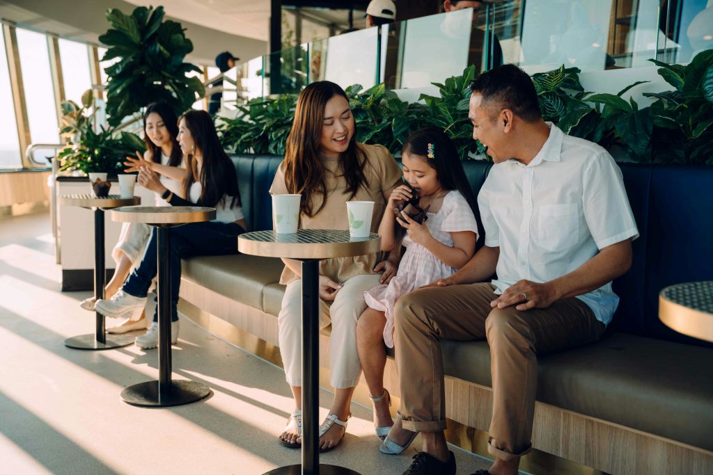 3인 가족과 2인 가족이 벤치에 앉아 녹음이 우거진 카페에서 음료를 마시고, 어른들이 미소를 짓는 동안 아이가 장난감을 가지고 놀고 있습니다.