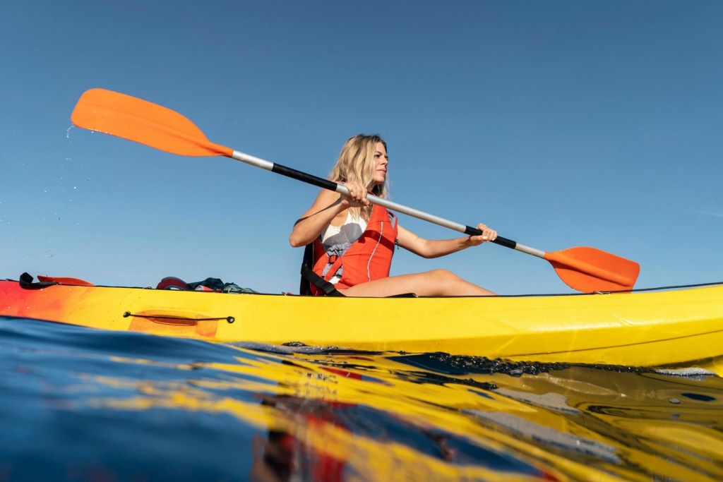 맑고 푸른 하늘 아래 잔잔한 물가에서 노를 저으며 빨간 구명조끼를 입은 노란색 카약을 탄 사람이 있습니다.