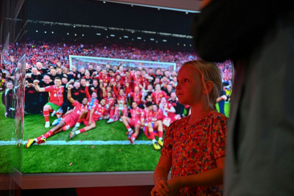 한 어린 소녀가 커다란 전광판 앞에 서서 팬들과 함께 경기장에서 축구팀을 축하하고 있습니다.