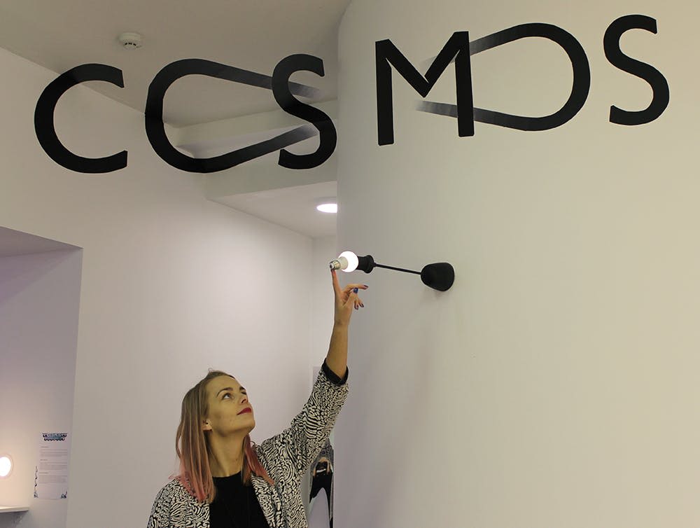 현대적인 인테리어와 벽에 '코스모' 라고 쓰인 커다란 검은 글자가 있는 방에서 한 여성이 벽에 불을 붙인 스콘스를 만지기 위해 손을 뻗습니다.