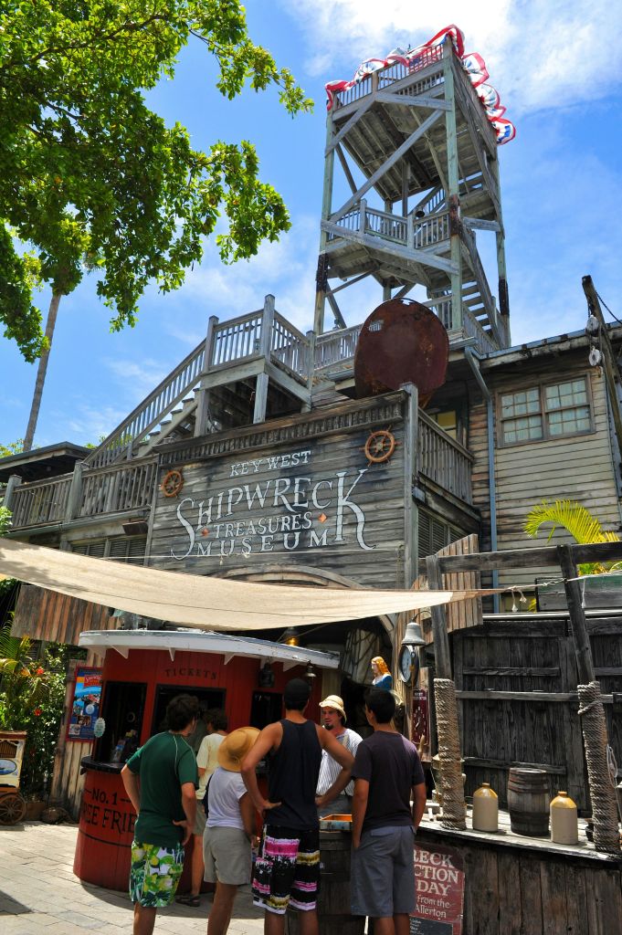 관광객들은 키 웨스트 난파선 보물 박물관 입구 밖에 서 있습니다. 키 웨스트 난파선 보물 박물관은 높은 탑과 해양 장식이 있는 목조 건물입니다.