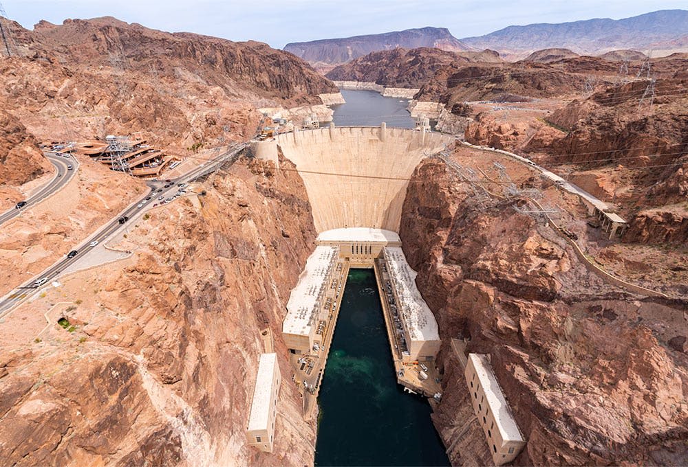 바위 지형으로 둘러싸인 커다란 저수지를 견디고 있는 후버 댐의 공중 전망, 인근에 건물과 도로가 보입니다.