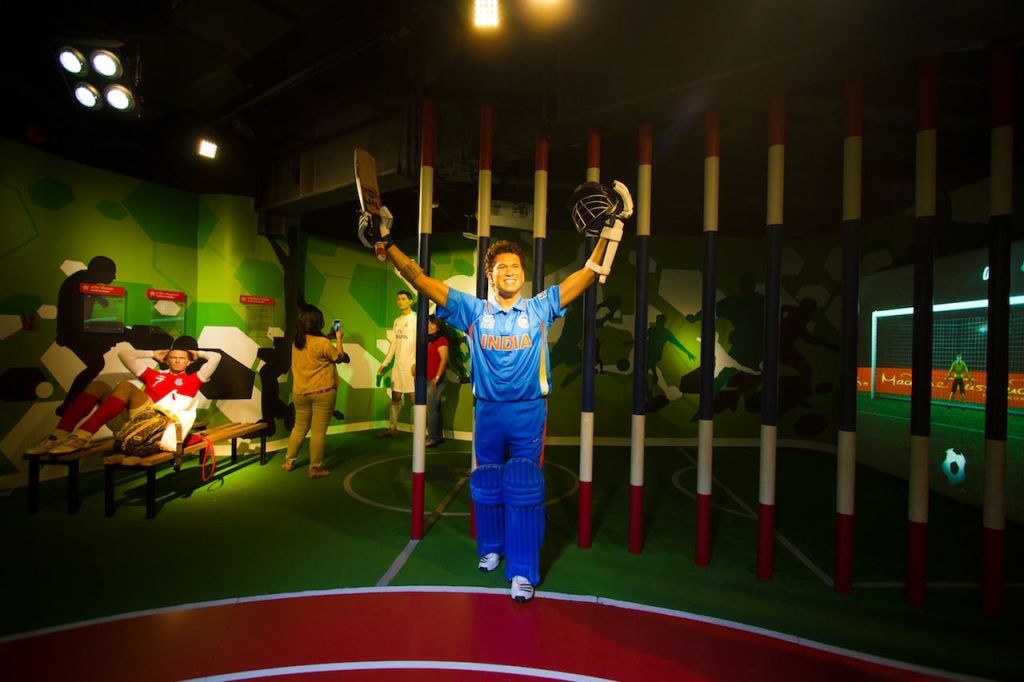 파란색 유니폼을 입은 인도 크리켓 선수의 왁스 형상이 실내 디스플레이에 박쥐와 헬멧을 들고 팔을 들어 올린 채 서 있습니다.