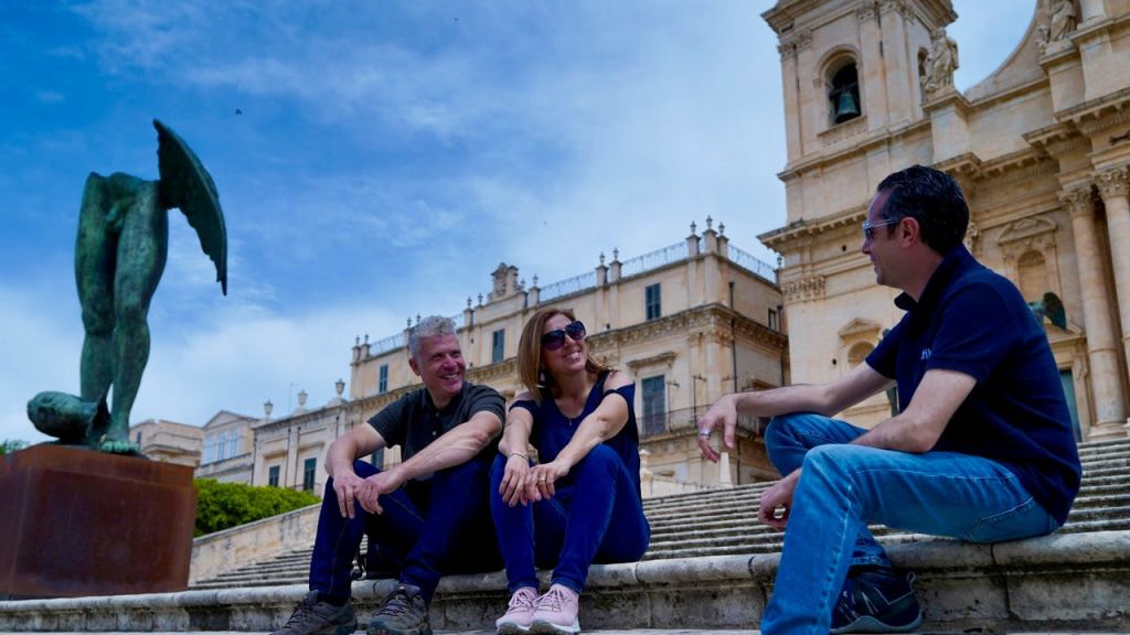 파란 하늘 아래 시계탑이 있는 유서 깊은 건물 앞 계단에 앉아 이야기를 나누는 세 사람.