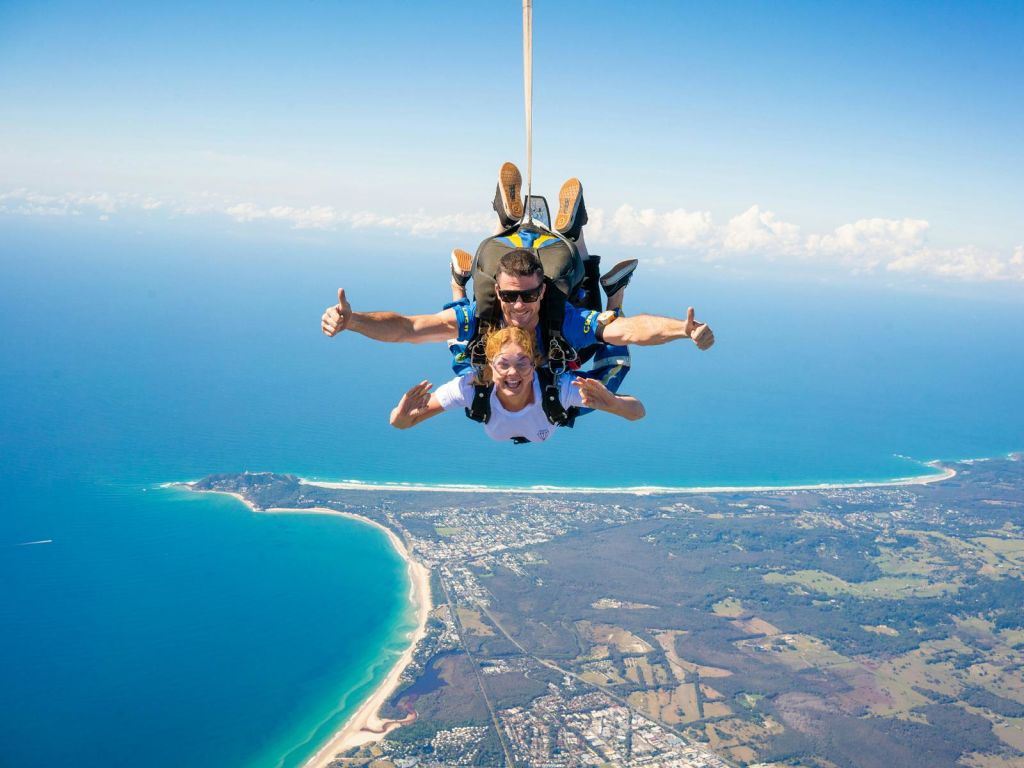 두 사람이 바다, 해안선, 육지가 보이는 해안 풍경을 보며 일렬로 스카이다이빙을 하며 미소를 짓고 엄지손가락을 치켜들고 있습니다.