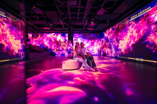 벽과 바닥에 보라색과 분홍색 디지털 투영이 있는 방의 벤치에 두 사람이 앉아 있습니다.