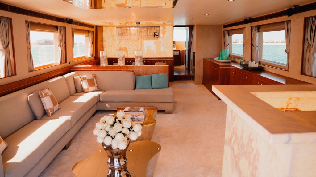 베이지색 단면 소파, 흰색 꽃이 놓인 유리 커피 테이블, 물이 내려다보이는 커다란 창문이 있는 고급 보트 거실 공간.