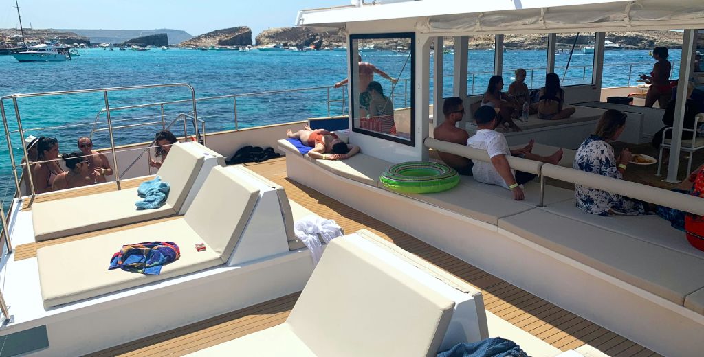 보트 갑판에서 휴식을 취하는 사람들도 있고, 그늘진 곳에 앉아 일광욕을 즐기는 사람들도 있습니다. 바다와 바위 해안선이 배경입니다.