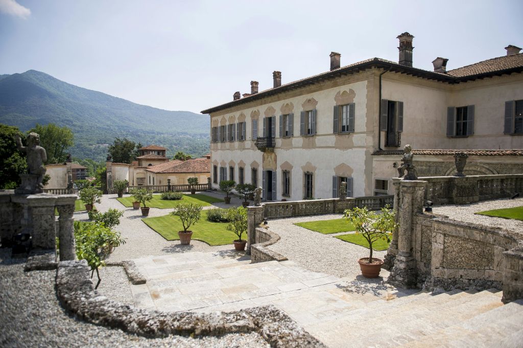 화창한 날 산악 지대를 배경으로 베이지색 벽, 녹색 셔터, 잘 다듬어진 정원이 있는 유서 깊은 저택입니다.