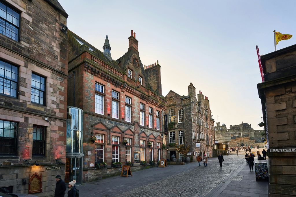 유서 깊은 석조 건물이 자갈길에 늘어서 있으며, 보행자가 몇 명 걸어가고 멀리 보이는 성이 있습니다.