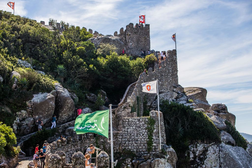 돌로 만든 성이 깃발과 관광객들이 흩어져 있는 언덕 꼭대기에 있습니다. 빽빽한 녹색 단풍이 밝은 하늘 아래 구조물을 둘러싸고 있습니다.