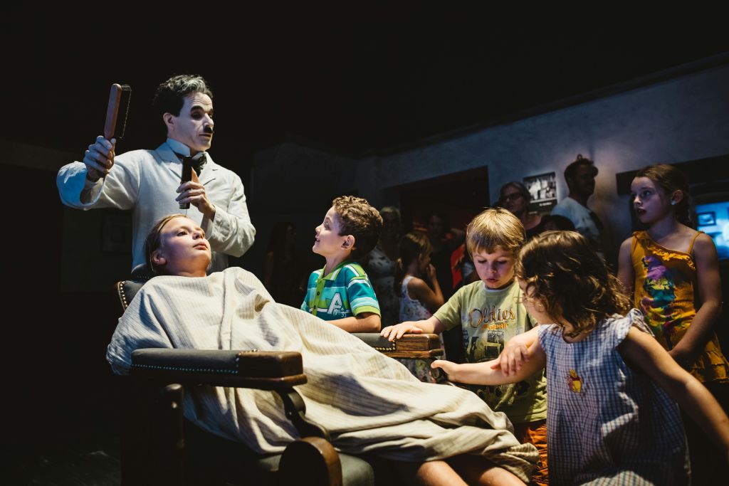 흰색 메이크업과 검은색 복장을 한 광대나 연기자를 닮은 인물이 시트로 덮인 사람 주위에 아이들이 모여듭니다.