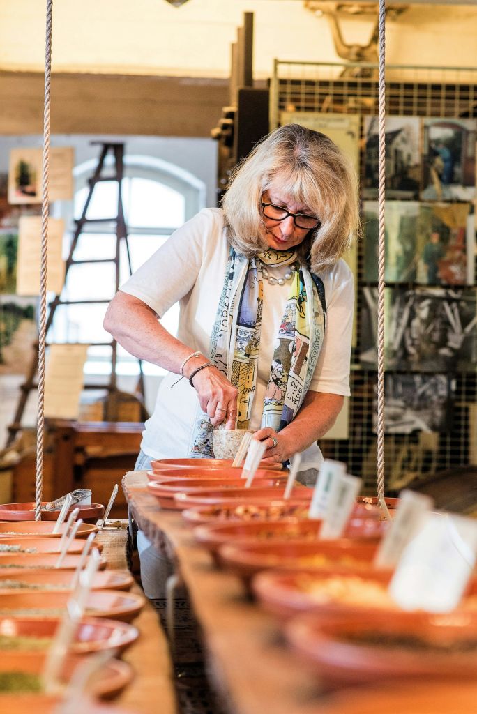 금발 머리에 안경을 쓴 여성이 예술 작품과 창문을 배경으로 예술 스튜디오에서 점토 작업을 하고 있습니다.