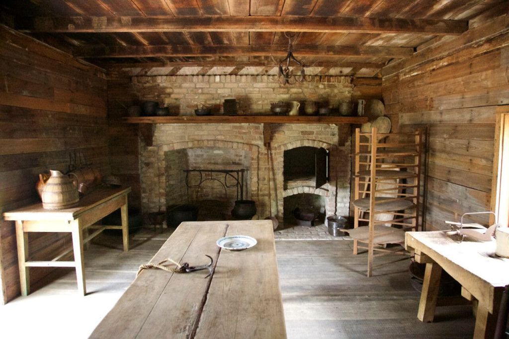 나무 테이블, 벽돌 벽난로, 냄비와 프라이팬이 있는 선반, 나무 건조대가 있는 소박한 주방.