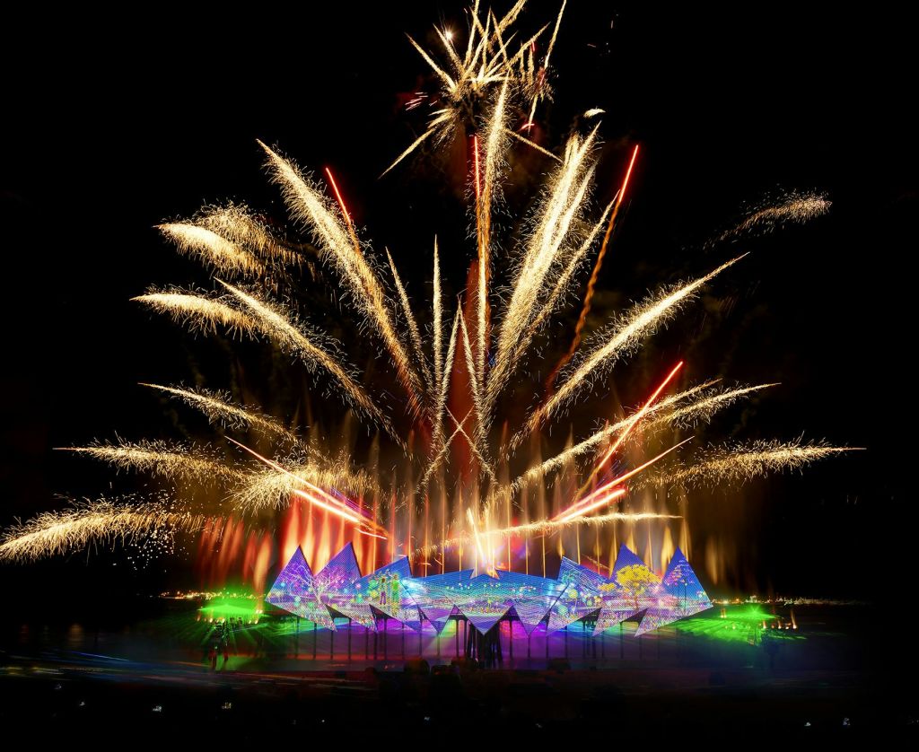 어두운 밤하늘을 배경으로 추상적인 모양의 화려한 무대 위로 밝은 불꽃놀이가 펼쳐집니다.