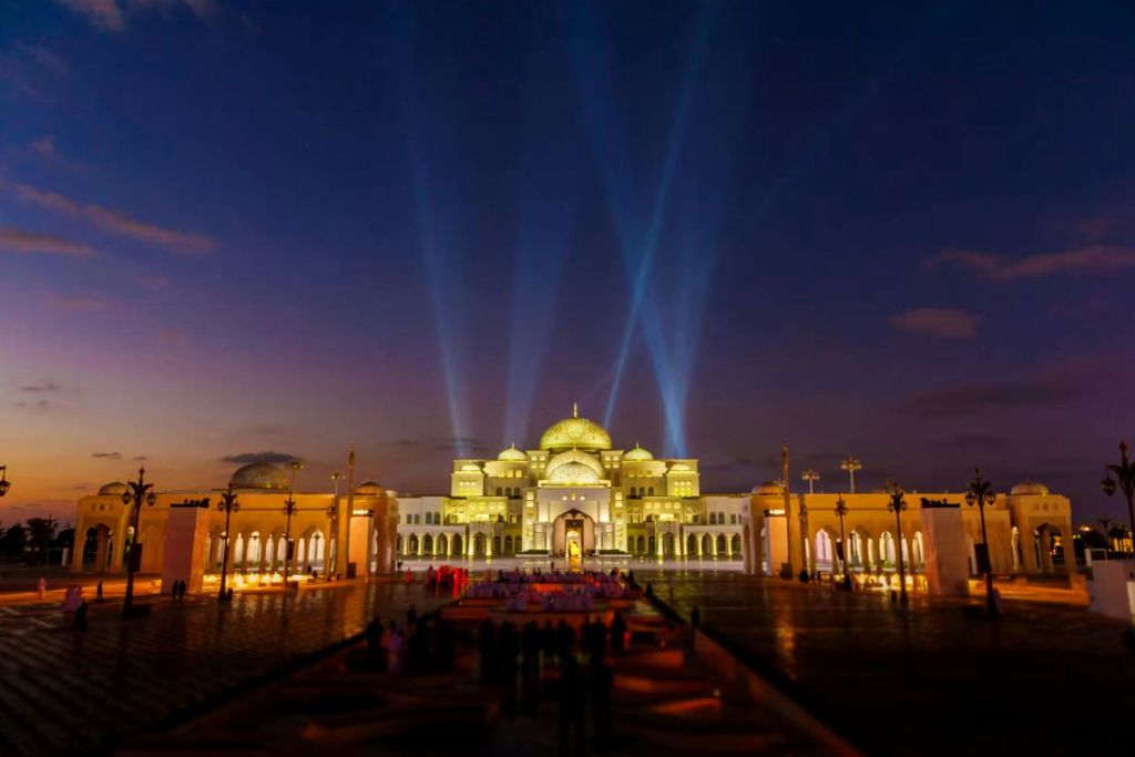 밤에는 돔과 아치가 있는 조명이 있는 궁전, 하늘로 투사되는 빛줄기, 앞에는 군중이 있습니다.