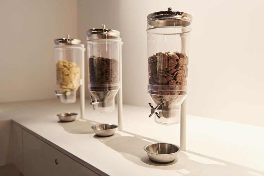 각각 아래에 금속 그릇이 있는 서로 다른 시리얼이 들어 있는 세 개의 원통형 디스펜서가 흰색 카운터에 놓여 있습니다.