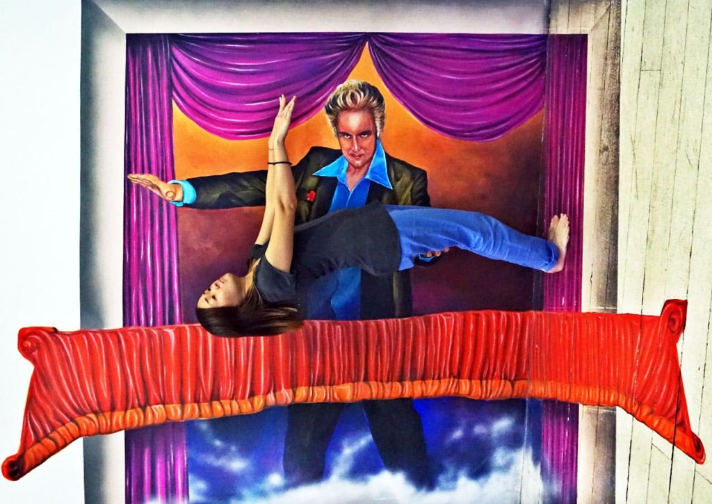 보라색 커튼 앞에 놓인 마술사를 배경으로 한 여성이 공중에서 떠다니는 듯한 수평 포즈를 취하고 있습니다.