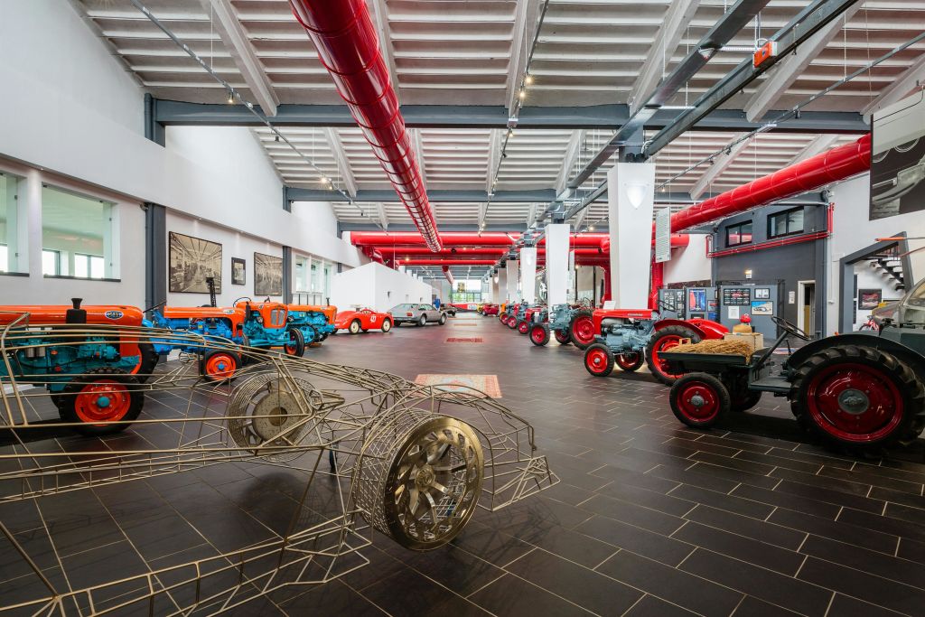 빈티지 트랙터와 와이어프레임 자동차 모델을 전시하는 넓은 실내 전시관으로, 빨간색 천장 파이프와 커다란 창문이 있습니다.