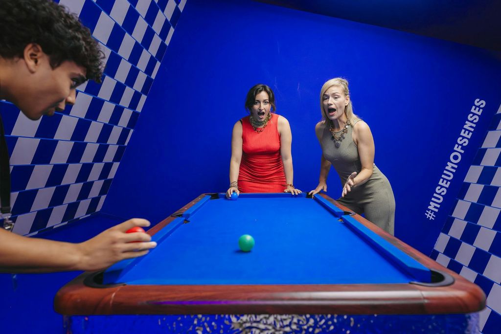 '감각의 박물관' 내부의 푸른색 당구대 주위에 녹색 공 하나가 놓인 두 여성이 깜짝 놀라며 서 있습니다.
