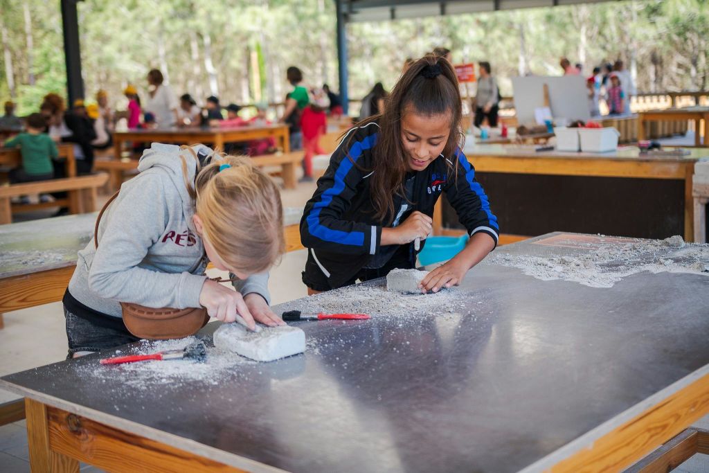 실습 활동에 참여하는 어린이들은 도구를 사용하여 야외 덮개 공간의 큰 테이블에 흰색 블록을 조각합니다.