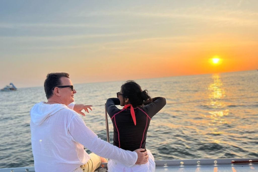 해 질 녘 보트에 앉아 있는 한 남녀의 두 팔이 레일에 얹혀 있고 멀리 떨어진 보트가 수평선에 보입니다.