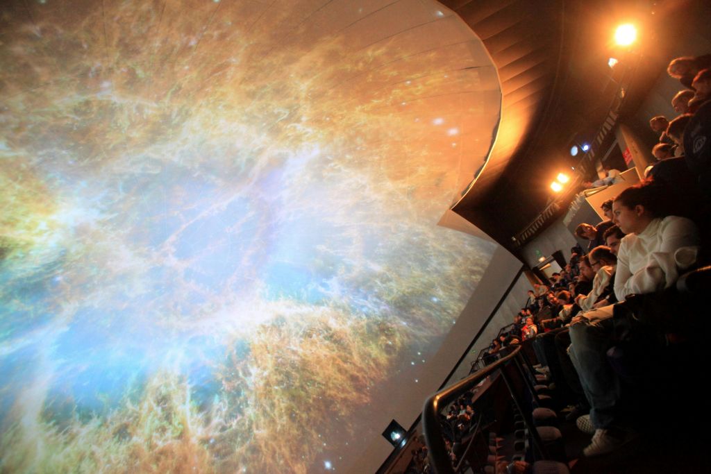 천문관에 앉아 커다란 돔형 천장에 우주의 성운이 투영된 광경을 지켜보는 사람들.