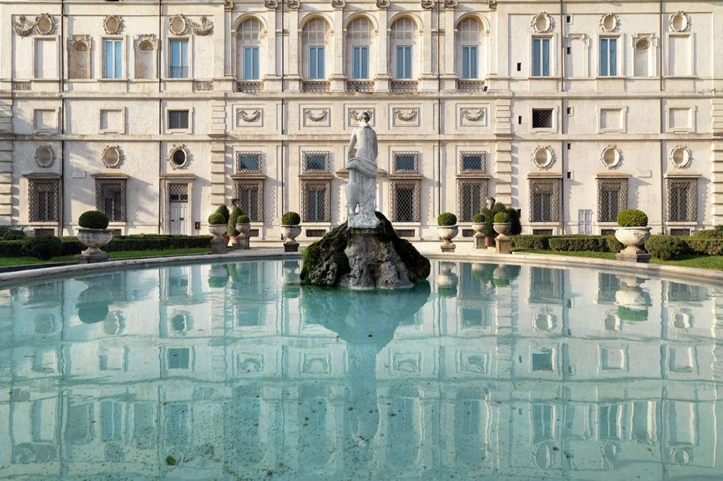 화려하고 고전적인 스타일의 건물 앞에 있는 원형 분수 중앙에 자리한 동상으로, 양쪽에 잘 손질된 덤불이 있습니다.