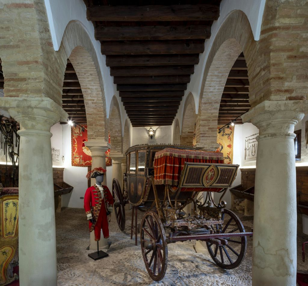 태피스트리로 둘러싸인 아치형 석조 천장 아래 마차가 있으며, 근처에 마침표 의상을 입은 마네킹이 있습니다.
