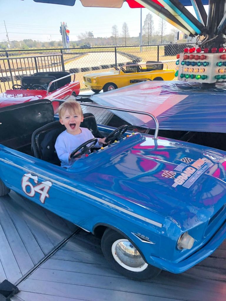 놀이공원에서 64번과 함께 파란색 승용차에 앉아 웃고 있는 어린 아이. 다채로운 색상의 차량이 눈에 띕니다.