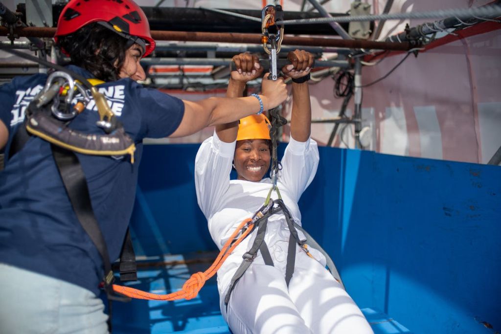 파란색 셔츠와 빨간색 헬멧을 착용한 다른 사람이 흰색 기어를 착용한 채 집라인에 고정된 채 미소를 짓고 있습니다.