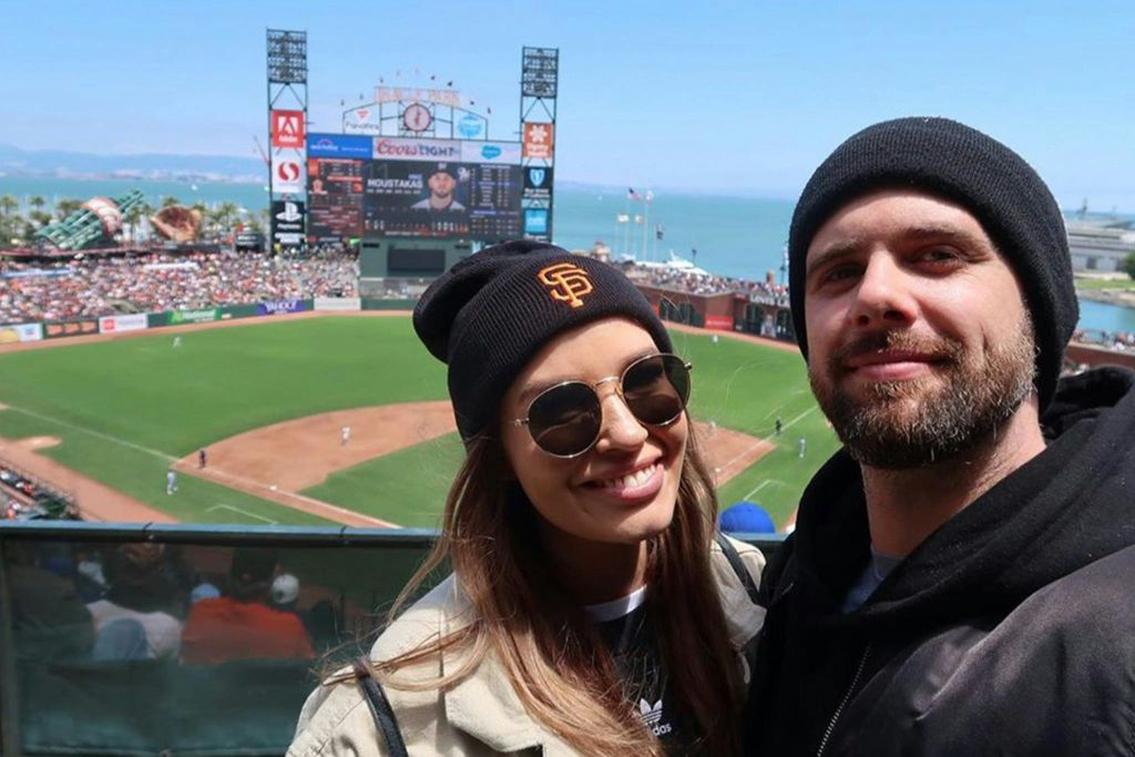 비니를 쓴 남녀가 경기장과 바다를 배경으로 야구장 앞에서 미소를 짓고 있습니다.