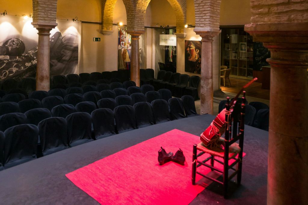 검은색으로 덮인 의자가 줄지어 무대와 마주보고 있는 소극장. 무대에는 레드 카펫과 소품이 놓인 나무 의자가 있습니다.