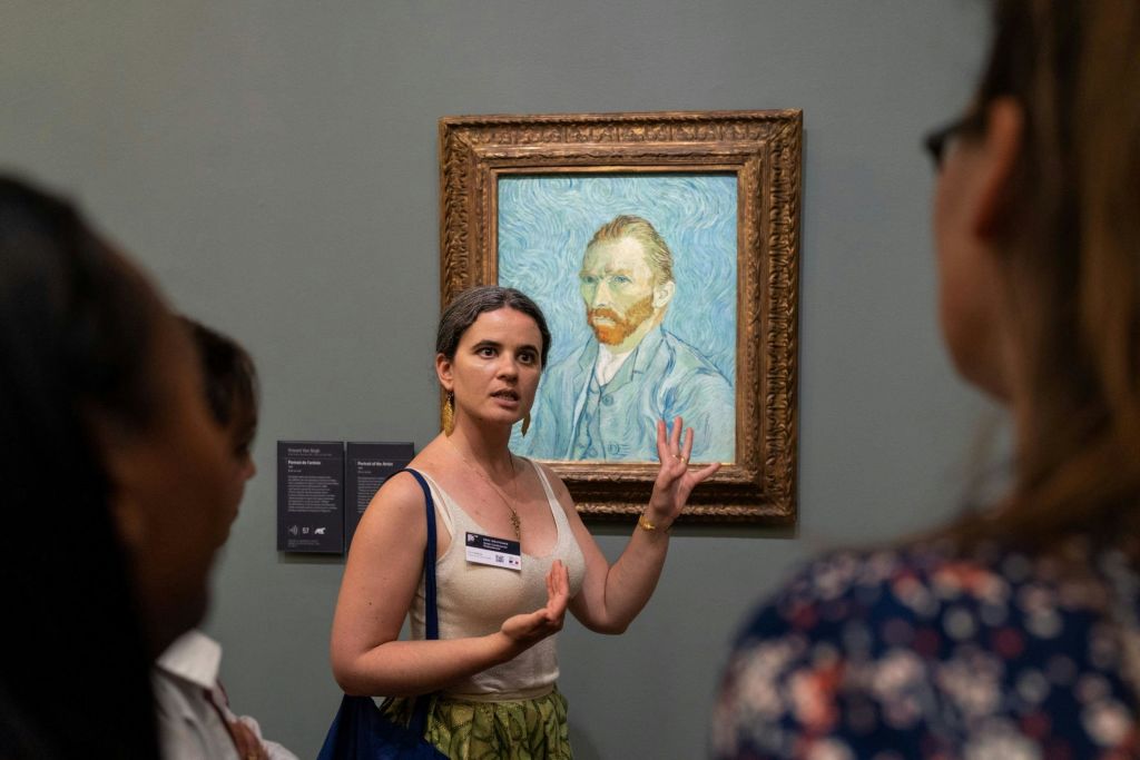 박물관에서 붉은 수염과 파란색 배경을 가진 남자의 액자 그림 근처에서 제스처를 취하는 사람.