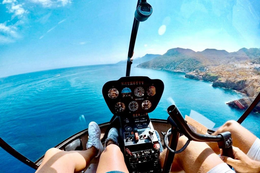 헬리콥터 조종석에서 바라보는 풍경으로, 바다와 해안선을 배경으로 앉은 두 사람의 조종석과 다리를 볼 수 있습니다.