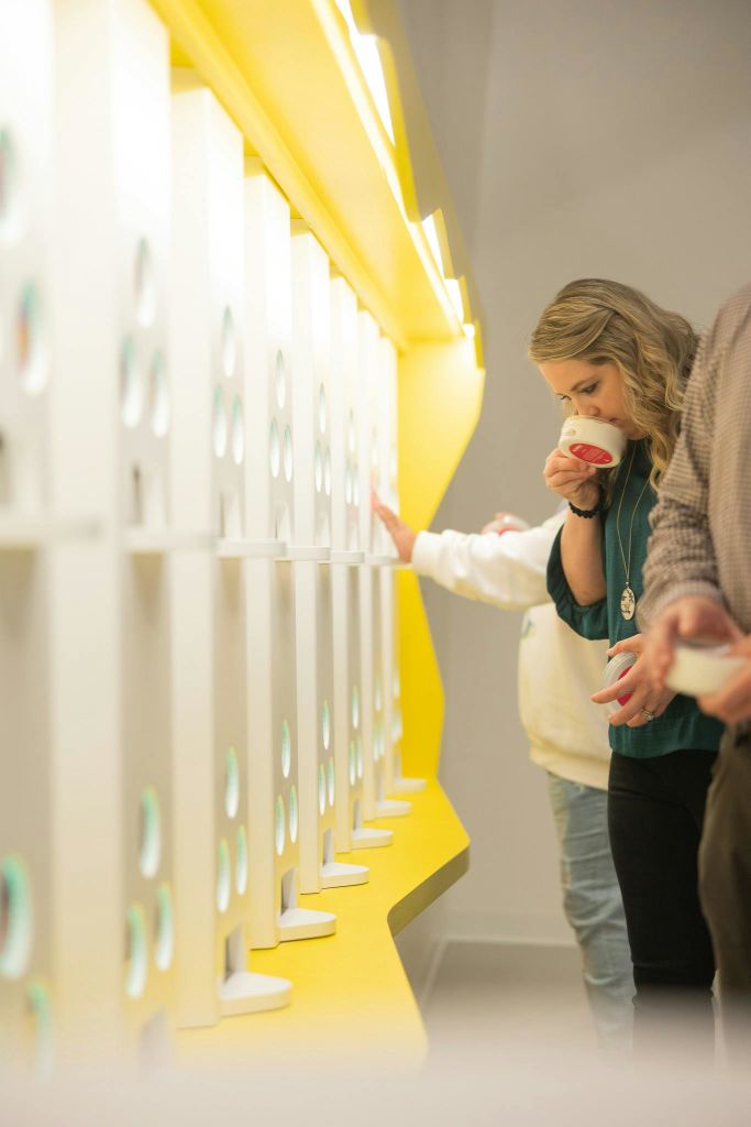 한 여성이 컷아웃과 노란색 조명이 있는 흰색 선반 옆에 서서 컵에서 음료를 마시고 있습니다. 다른 두 사람이 부분적으로 보입니다.