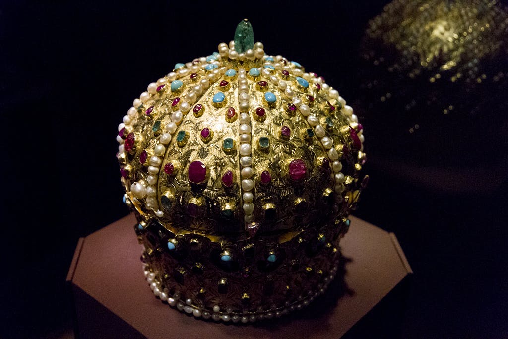 진주, 루비, 청록색, 에메랄드로 장식된 화려한 황금 왕관이 받침대에 전시되어 있습니다.