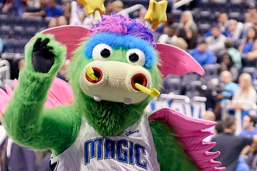 스포츠 행사에서 커다란 눈과 날개를 가진 녹색의 다색 마스코트이며, 배경은 흐릿한 관중석입니다.