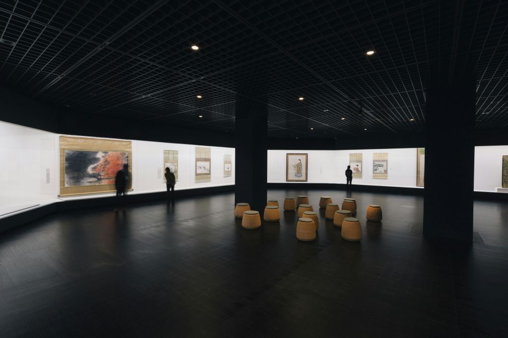 흰색 벽에 액자에 담긴 예술품으로 조명이 희미한 아트 갤러리를 꾸며보세요. 두 사람이 스툴이 중앙에 배치된 상태에서 예술을 관찰합니다.