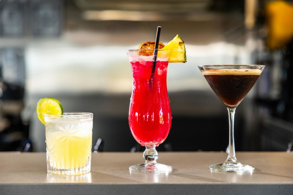 카운터에 세 가지 칵테일: 라임이 들어간 노란색 음료, 파인애플 장식이 들어간 빨간색 음료, 마티니 잔에 담긴 진한 음료.