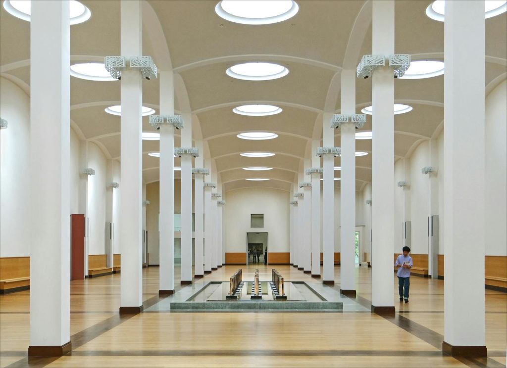 높은 아치형 천장, 채광창, 줄지어 있는 기둥이 있는 넓은 객실. 한 사람이 직사각형 분수대 근처에 서 있습니다.