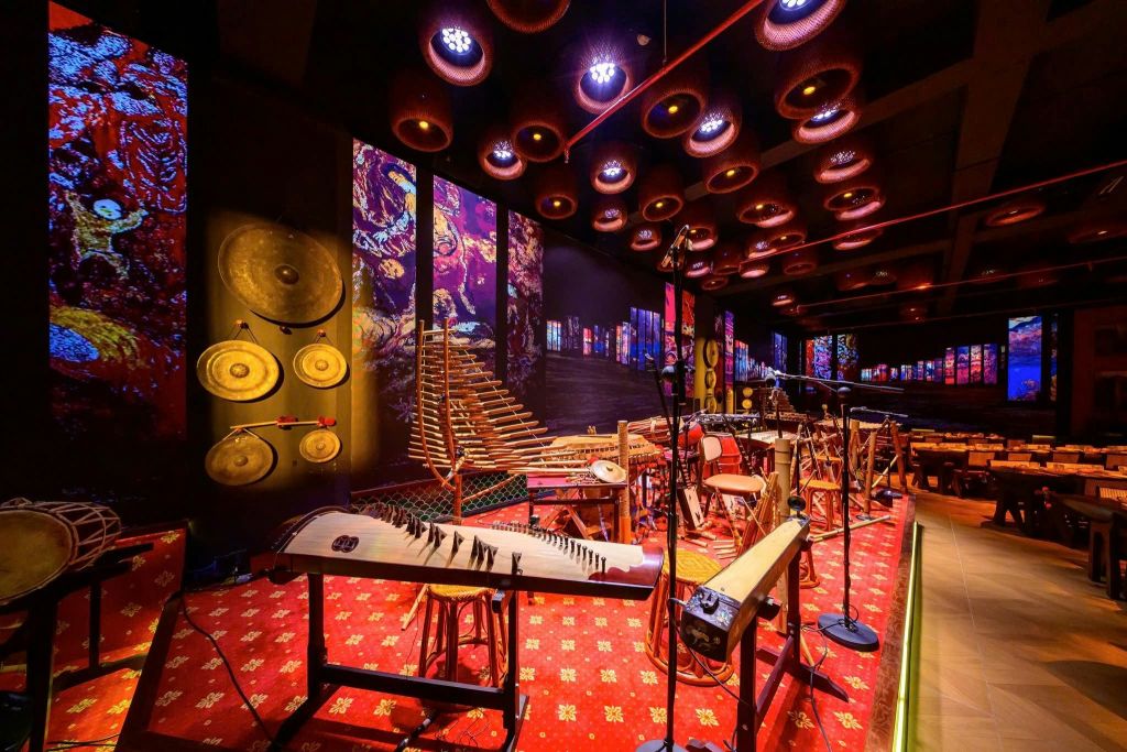 조명이 켜진 원형 천장 조명 아래 전통 악기, 생동감 넘치는 벽면 예술품, 무늬가 있는 레드 카펫이 가득한 객실입니다.