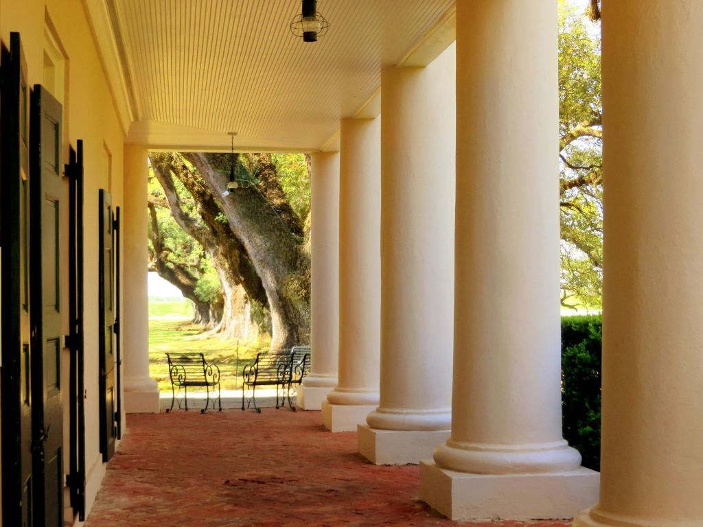 철제 의자, 벽돌 바닥, 커다란 나무를 배경으로 한 베란다의 흰색 기둥.