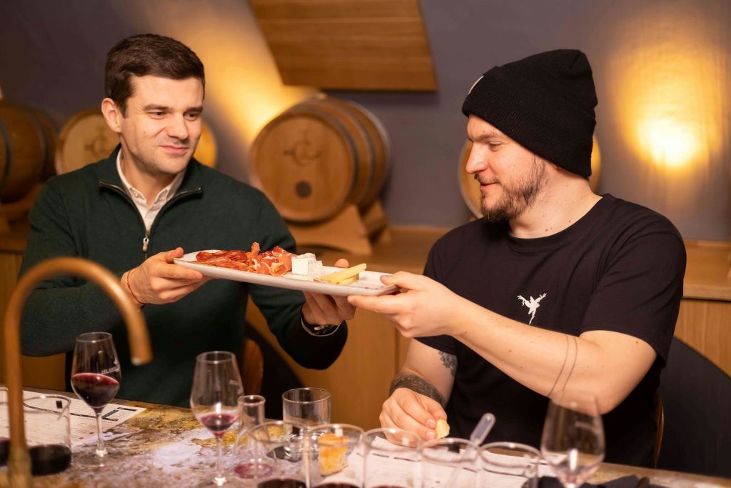 와인잔과 통을 배경으로 모듬 샤퀴트리 접시를 지나고 있는 두 남자.