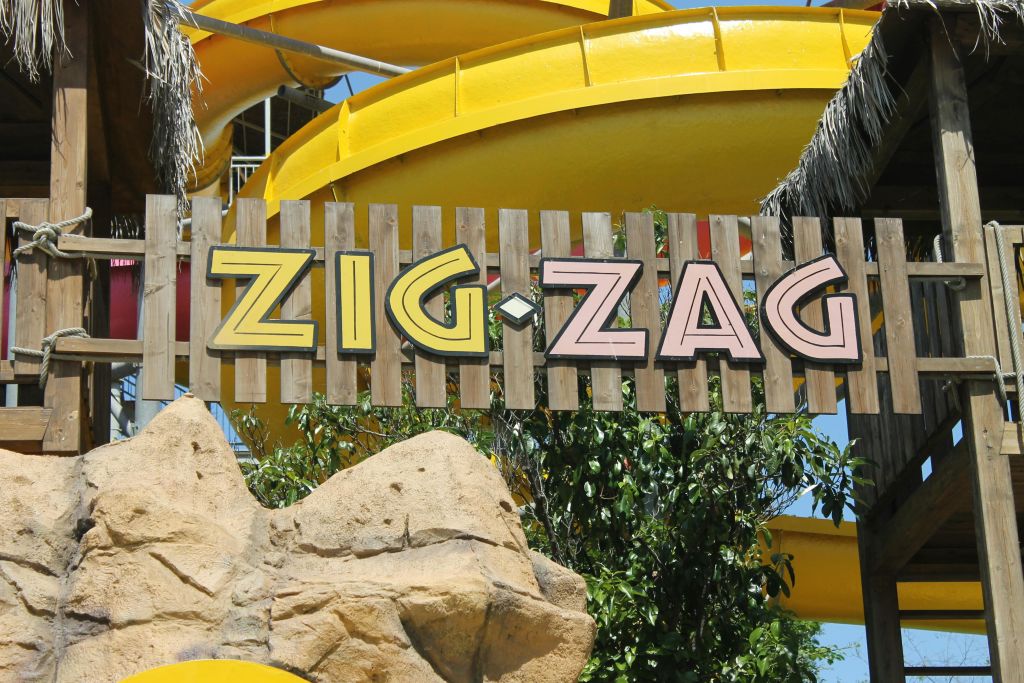 노란 물 미끄럼틀, 바위, 녹지 앞에 화려한 글씨로 "지그재그" 가 있는 나무 간판.
