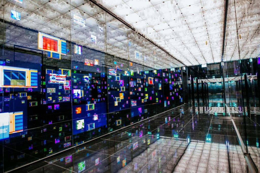 반사되는 검은색 바닥, 다채로운 디지털 화면을 보여주는 거울 벽, 밝은 천장이 있는 미래형 복도.