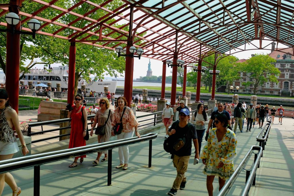 자유의 여신상을 배경으로 해변 근처의 그늘진 길을 걷는 사람들.