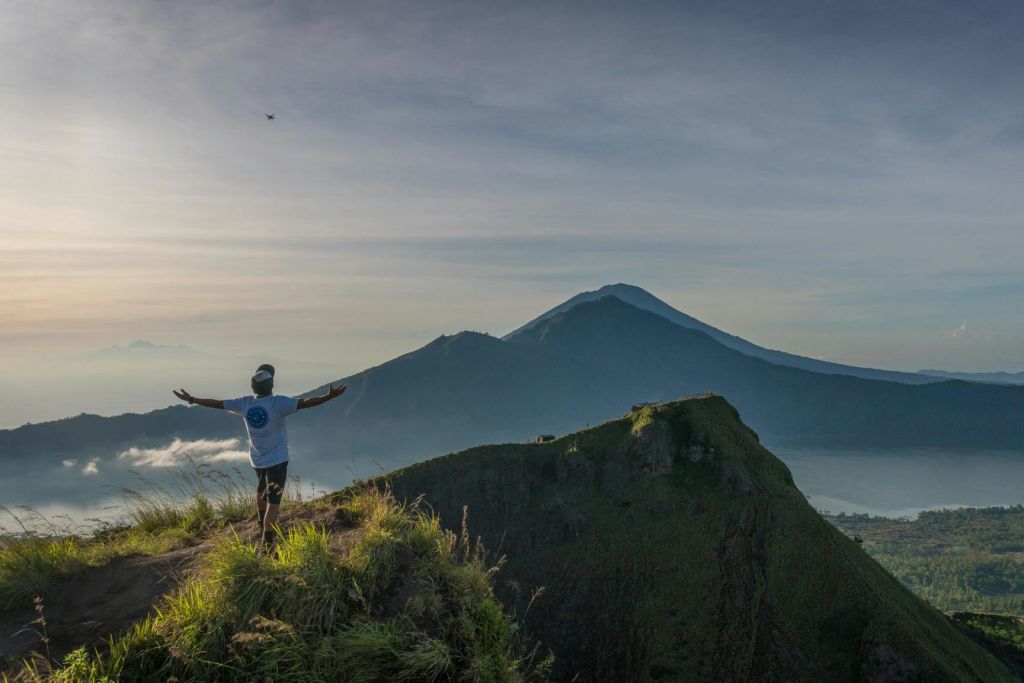 한 사람이 양팔을 쭉 뻗은 채 풀이 무성한 산봉우리에 서서 맑은 하늘 아래 먼 산을 마주하고 있습니다.